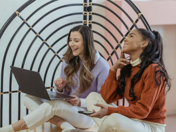 Two women laughing as the work on a laptop and a phone to illustrate creating social media campaigns